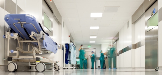 corridoio di un ospedale dove si intravedono lontano dei medici in camice verde. in primo piano, un letto ospedaliero